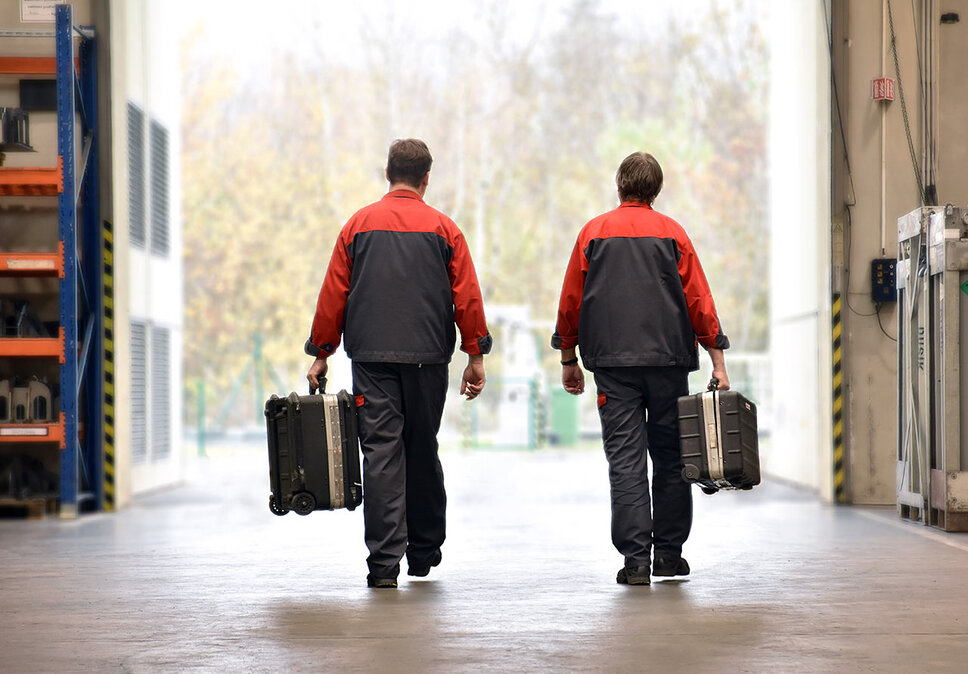 Zwei Werkzeugbautechniker, ausgerüstet für einen Service-Einsatz, gehen in Richtung eines offenen großen Tores. Sie tragen jeweils einen Werkzeugkoffer, der für ihre bevorstehende Aufgabe notwendig ist. Die Szene vermittelt eine Atmosphäre von Professionalität und Bereitschaft, mit Fokus auf die Techniker und ihre Ausrüstung.