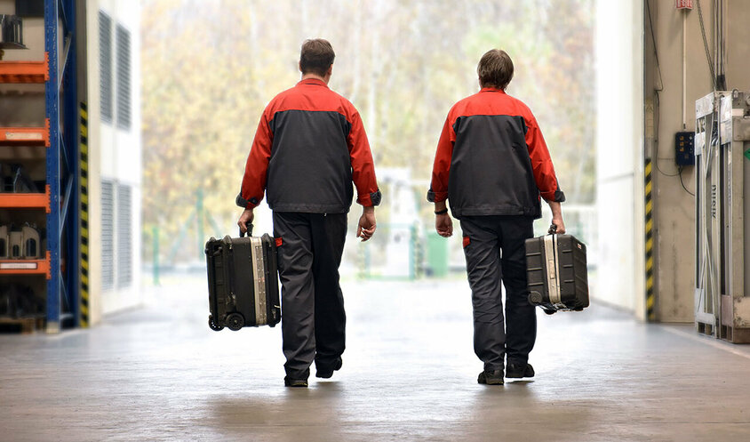Two employees from weba's mobile tool manufacturing service are seen in work attire, carrying toolboxes as they navigate a workshop. They are on their way to install or maintain forming and stamping tools on-site at a customer location.
