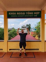 Jan Wengryn standing under a sign reading “General Surgery Department” in Vietnam, smiling and pointing upward – a light-hearted moment from his journey.