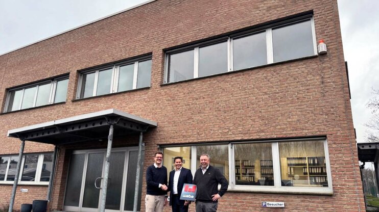 Three men standing in front of a brick building, two shaking hands while the third holds a document with weba branding.