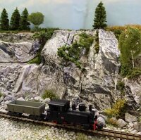 Černá modelová parní lokomotiva s nákladními vozy před detailně zpracovanou skalnatou krajinou.