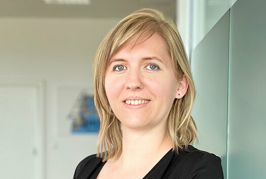 Portrait of Tanja Burghuber, HR Manager at weba Werkzeugbau, leaning casually against a frosted glass wall in Dietach, with a professional and friendly expression, highlighted by soft lighting and a blurred background. The photo is used in the contact section of the job openings for the Steyr location of weba Werkzeugbau.