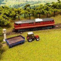 Detailní záběr na modelovou železnici s červenou lokomotivou, traktorem a přívěsem v upravené krajině.