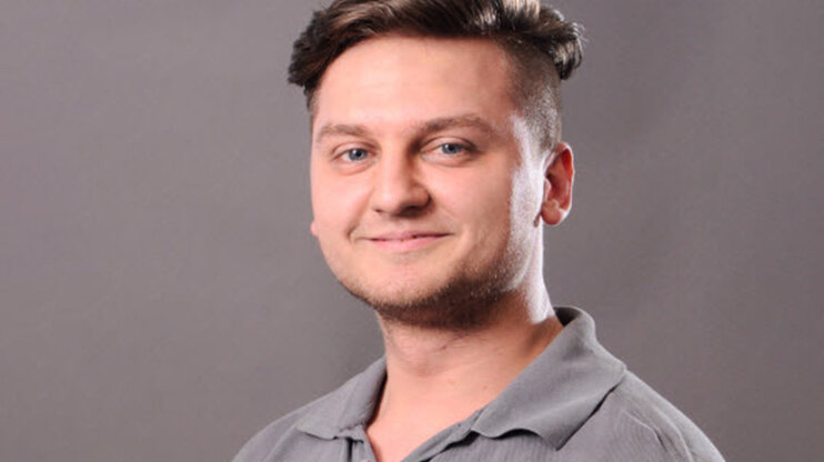 Portrait of a young man with short, side-trimmed hair and a friendly smile. He is wearing a gray polo shirt and looking directly at the camera. The background is neutral and gray.