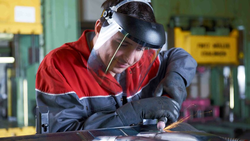 weba Mitarbeiter bearbeitet ein Metallbauteil mit einem Schleifwerkzeug und trägt dabei Gesichtsschutz und Schutzhandschuhe.